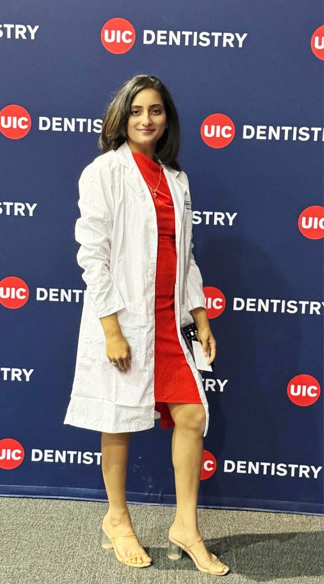 Shahana in a white coat and red dress in front of a UIC Dentistry backdrop